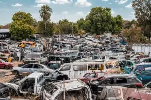 A busy junkyard filled with a variety of old cars under a clear sky, surrounded by trees.